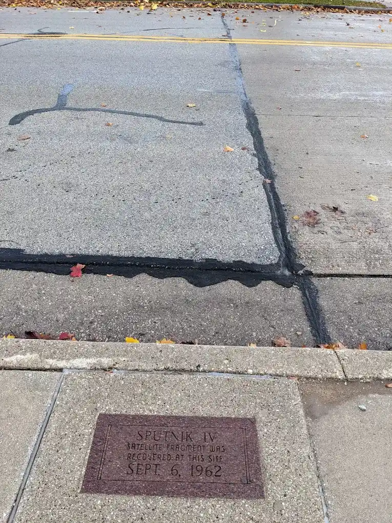 Commemorative sign on the sidewalk, adjacent to the spot where the fragment of Sputnik IV was recovered, a site designated by a metal hoop in the asphalt.