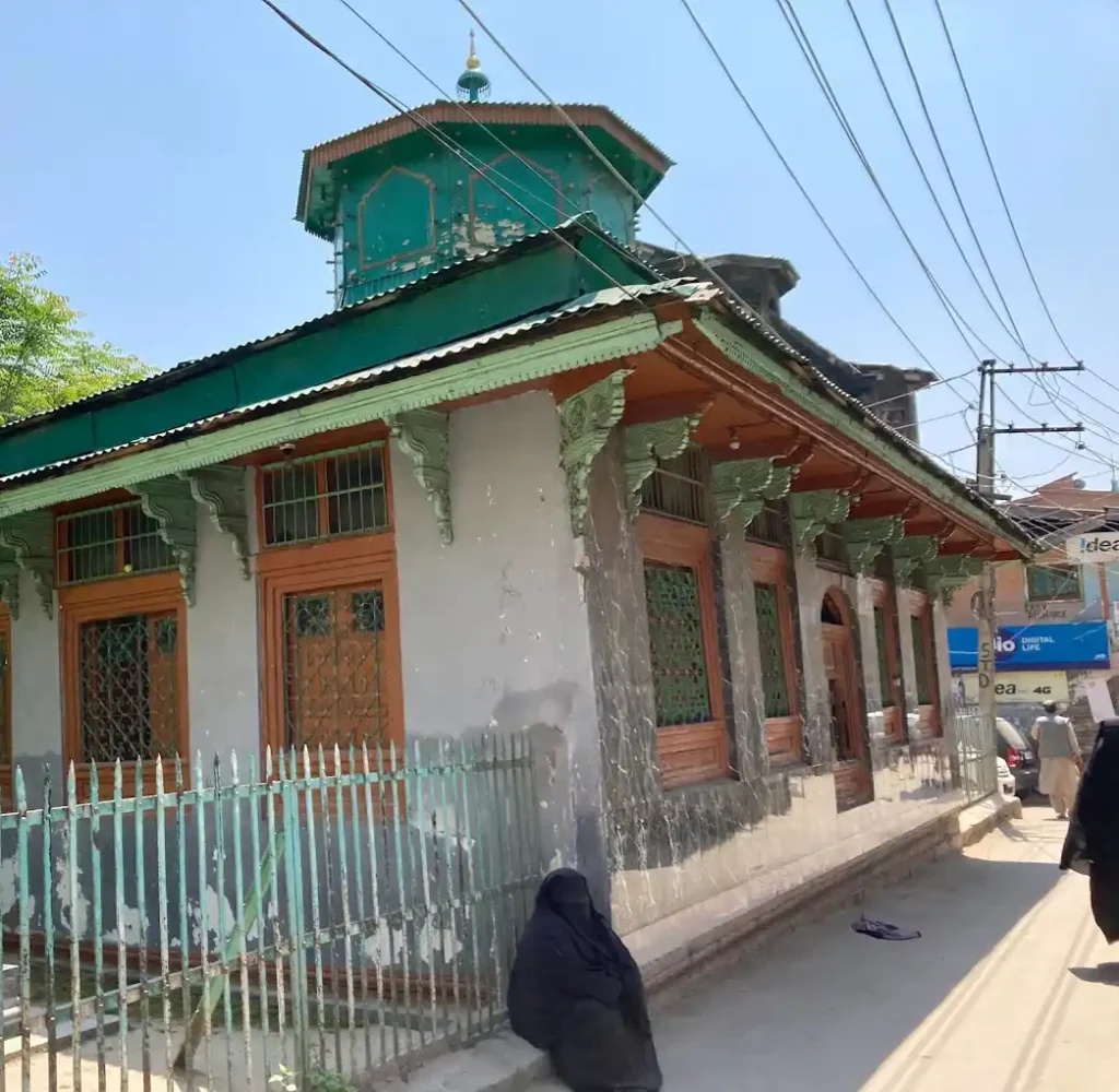 A woman in a burka sits silently outside the Roza Bal shrine in Srinagar, while another woman walks by in the background.